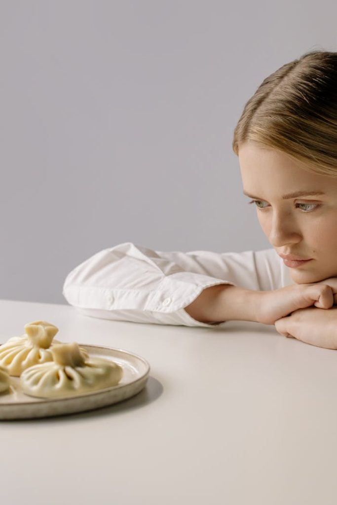 Dlaczego jedzenie budzi poczucie winy? Jak przestać karać się za to, co masz na talerzu. A woman in a white shirt thoughtfully gazes at a plate of dumplings indoors.