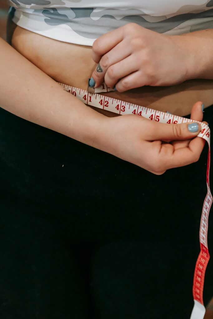 Oporność na odchudzanie, przewlekły stres, Traumę i Teorię Poliwagalną (psychosomatykę wagi) High angle of crop faceless overweight female measuring waist with centimeter for weight loss